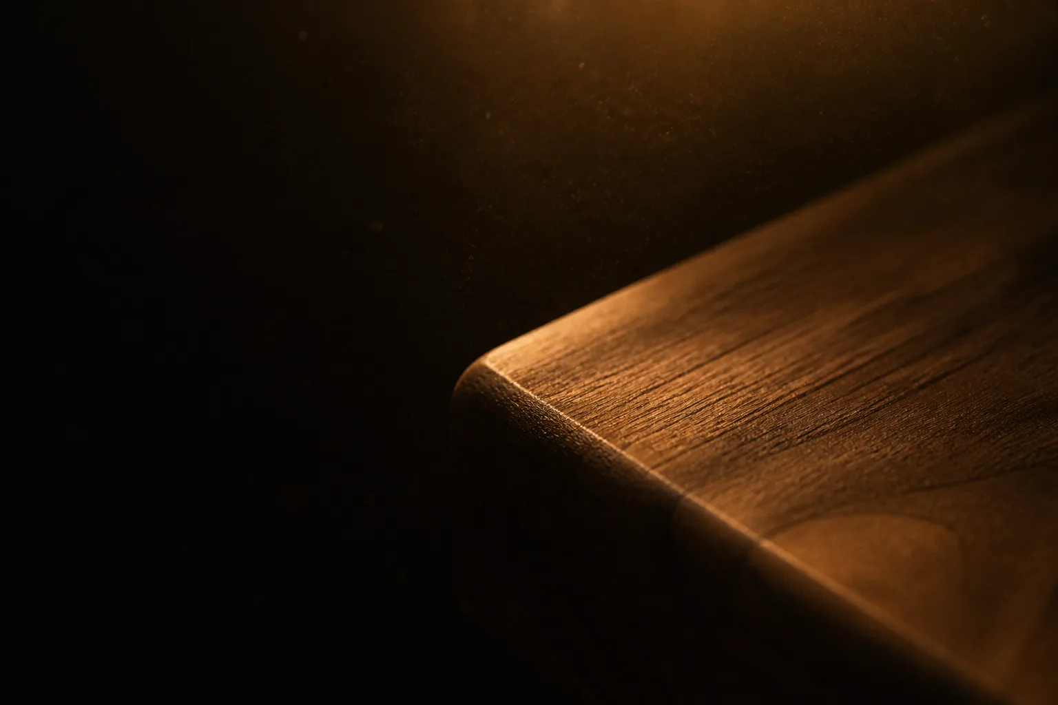 Low amber light grazing across the softly rounded edge of a dark timber bench top, the rest of the frame falling away into shadow.